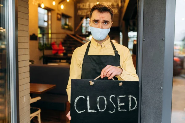 Sad waiter, barista or small business owner. Unhappy man in a medical protective mask and a black work apron stands at the entrance to a cafe, restaurant or bar and holds a CLOSED sign