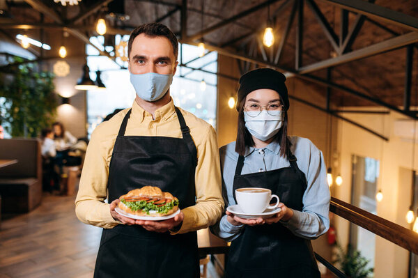 Friendly multiracial waiters wearing protective masks stand inside a restaurant, cafe or bar. The guy holds a fresh sandwich and the girl holds a cup of aromatic coffee