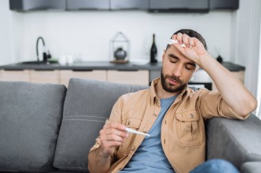 Mutsuz, hasta, beyaz bir adam kanepeye oturur sağlıksız hisseder, termometreye hayal kırıklığı içinde bakar, yüksek ateşi olan hasta bir adam, başı ağrır ve tedaviye ihtiyacı vardır, grip, karantina...