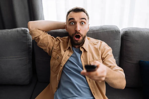 Shocked worried guy watch on tv sport channel, sitting on the couch holds a tv remote control, surprised with results of match. Emotional male nervous about game, football fan concept