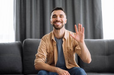 Video konferansı sırasında neşeli beyaz bir adamın portresi, kameraya bakıp el sallıyor ve gülümsüyor, arkadaşlarına, ailesine ya da meslektaşlarına selam veriyor, kanepede oturuyor.