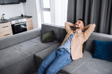 Tatmin olmuş, sakin, yakışıklı, beyaz adam kanepede gözleri kapalı oturuyor. Hafta sonları ya da tatil planları yapıyor ve gülümsüyor. Genç serbest çalışan çevrimiçi işten dinleniyor