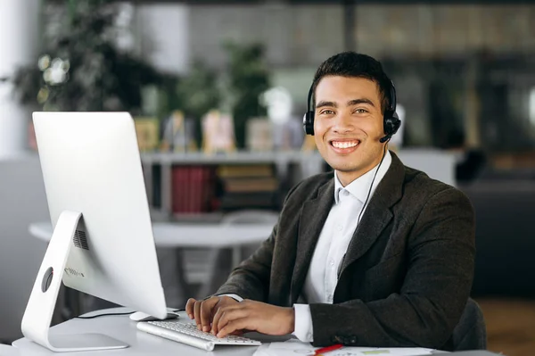 Support service, help desk. Portrait of a young successful hispanic call center worker, manager or entrepreneur sitting at work desk wearing headphones looking at the camera with a smile