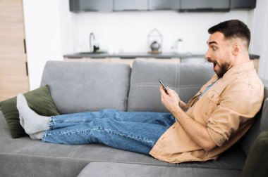 Aşırı neşeli Kafkasyalı adam kanepede oturuyor, akıllı telefon kullanıyor. Yakışıklı adamın hayallerindeki işi ya da iyi haberi ya da mesajı var. Çekici stil adamı arkadaşlarıyla ya da ailesiyle internette sohbet ediyor.