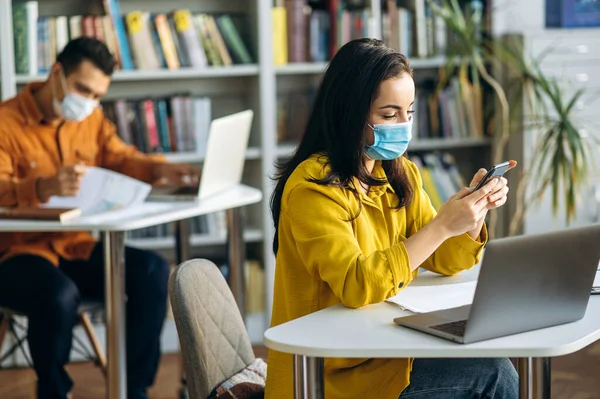 Learning during a pandemic. Students sit at a distance from each other with protective masks on their faces. A student girl sits at a desk during a lesson in a medical mask and chatting online