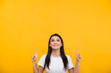 Beyaz tişörtlü, gülümseyen, mutlu, beyaz tenli, esmer bir kadın. Parmaklarını başının üstüne koy ve gülümse. Çalışma alanı, izole edilmiş turuncu arka plan.