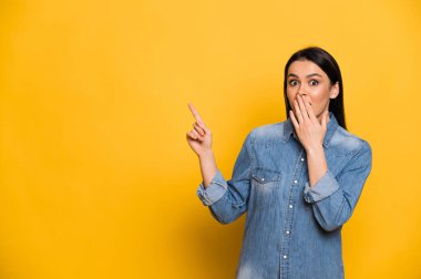 Heyecanlı, beyaz, mavi gömlekli güzel esmer kadın, ağzını eliyle kapatıyor ve şaşırıyor. Başparmağı yukarıda, izole edilmiş turuncu arka planda duruyor.