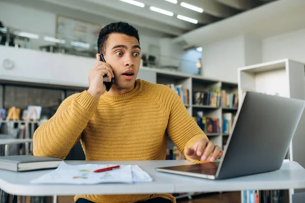 Hispanic businessman talking with colleague by smartphone, hearing shocked news. Young adult male employee looking at the laptop screen, receive unexpected message