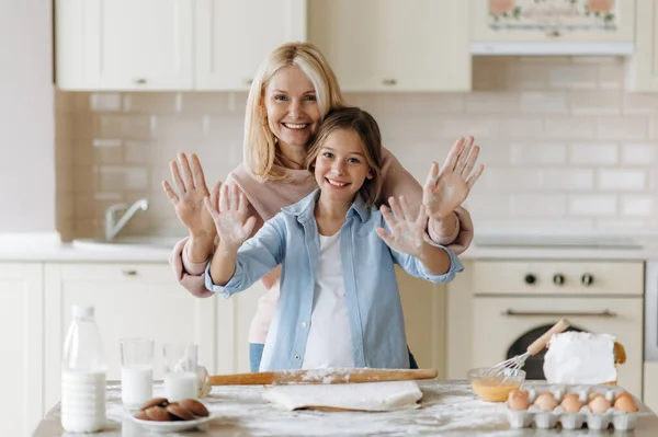 Caucasian happy mom and her beloved little daughter spend time together in the kitchen. Grandma teaches her cute granddaughter to cook pie, they are happy to cook together, having fun and laughing