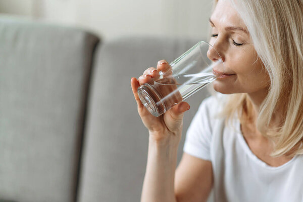 Следите за здоровым образом жизни. Крупным планом фотография зрелой кавказской привлекательной женщины, сидит на диване в гостиной, наслаждается питьем стакана чистой воды, ежедневной нормой