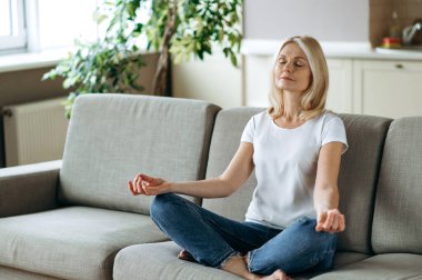 Tatmin olmuş orta yaşlı bir kadın evdeki kanepede oturuyor ve dinleniyor. Sarışın olgun, yoga ve meditasyon yapıyor. Lotus pozisyonunda, gözleri kapalı, stres giderici, sakinlik konsepti.