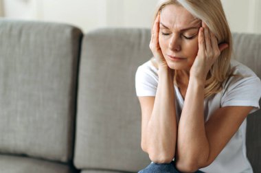 Baş ağrısı ya da stres. Orta yaşlı sarışın kadın oturma odasında ellerini başının üstüne koyup oturuyor. Baş ağrısı, stres, hastalık ya da kötü haber yüzünden mutsuz hissediyor. Dinlenmesi ve desteğe ihtiyacı var.