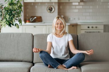 Tatmin olmuş orta yaşlı bir kadın evdeki kanepede oturuyor ve dinleniyor. Sarışın olgun, yoga ve meditasyon yapıyor. Lotus pozisyonunda, gözleri kapalı, stres giderici, sakinlik konsepti.