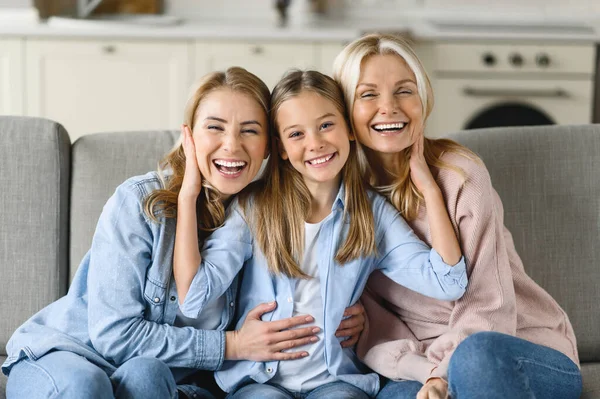 Three female generation. Happy caucasian grandmother, daughter and granddaughter are sitting at home on the couch, hugging, looking at the camera and smiling. Family relationships and values