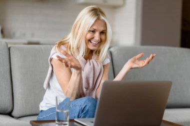 Oturma odasındaki kanepede oturan, bilgisayarını kullanan, uzaktan çalışan, iş arkadaşlarıyla iletişim kuran, mutlu, beyaz, son sınıf öğrencisi bir kadın.