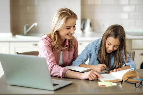 Learning at home. Caring mom helps her beloved daughter do her homework, they sit at the table at home, using a laptop, books and a notebook. Homeschooling