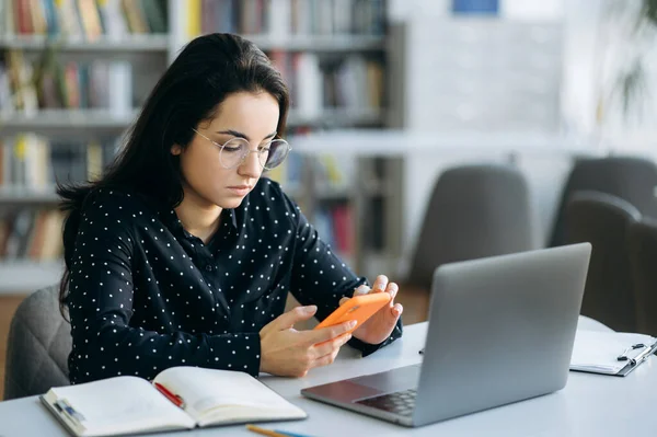 Serious young lady using smartphone, sit at the desk. Pretty woman in stylish wear browsing internet, chatting with colleagues or replies to an email