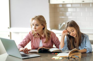 Mesafe öğrenme. Anne, kızına ödevlerini yapmasına yardım ediyor. Evde dizüstü bilgisayar, kitap ve defter kullanarak oturuyorlar. Evde eğitim