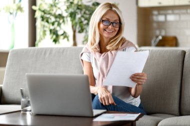 Orta yaşlı çekici bir sarışın kadın evde çalışıyor oturma odasındaki koltukta uzaktan çalışıyor, aylık raporları analiz ediyor, dizüstü bilgisayar kullanıyor. İnternette çalışmak ya da çalışmak