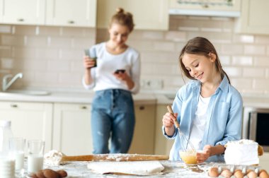 Annenin küçük yardımcısı. Beyaz tenli tatlı bir kız turta pişirmeyi öğreniyor, yumurta çırpıyor, gülümsüyor ve annesi arka planda telefonla duruyor.