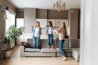 Neşeli beyaz anne, kız ve modern büyükanne evde birlikte eğleniyorlar, dans ediyorlar, kanepede zıplıyorlar, oynaşıyorlar ve gülüyorlar.