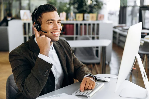 Joyful employee is consulting people, sitting in modern office. Successful hispanic business wearing headset man work as call center operator, communicate with clients, conducts online meeting