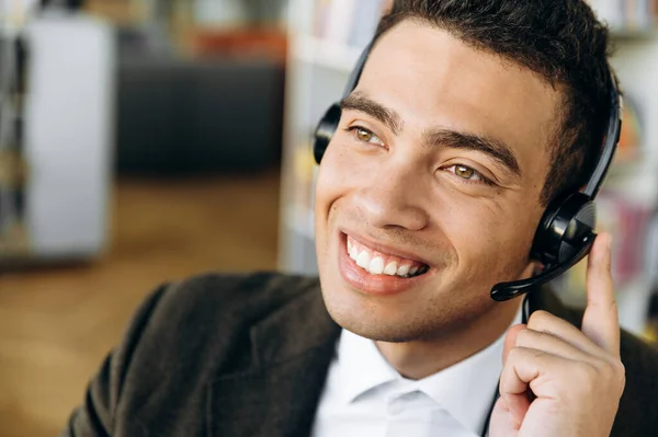 Close-up portrait of smiling hispanic male consultant, operator of call center sitting at the desk, wearing headset. Young adult employee is talking with people, support service concept