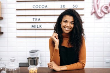 Üniforma giyen gülümseyen güzel bir garsonun portresi. Genç ve güzel bir Afro-Amerikan bayan barmen kameraya gülümsüyor, kafe tezgahının arkasında duruyor. Küçük işletme sahibi
