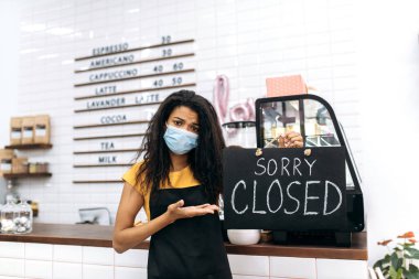 Üzgün Afrikalı Amerikalı garson, barmen ya da küçük işletme sahibi. Siyah önlüklü ve tıbbi maskeli mutsuz genç bir kadın kahve dükkanının, restoranın veya barın içinde duruyor ve elinde kapalı tabela levhası var.