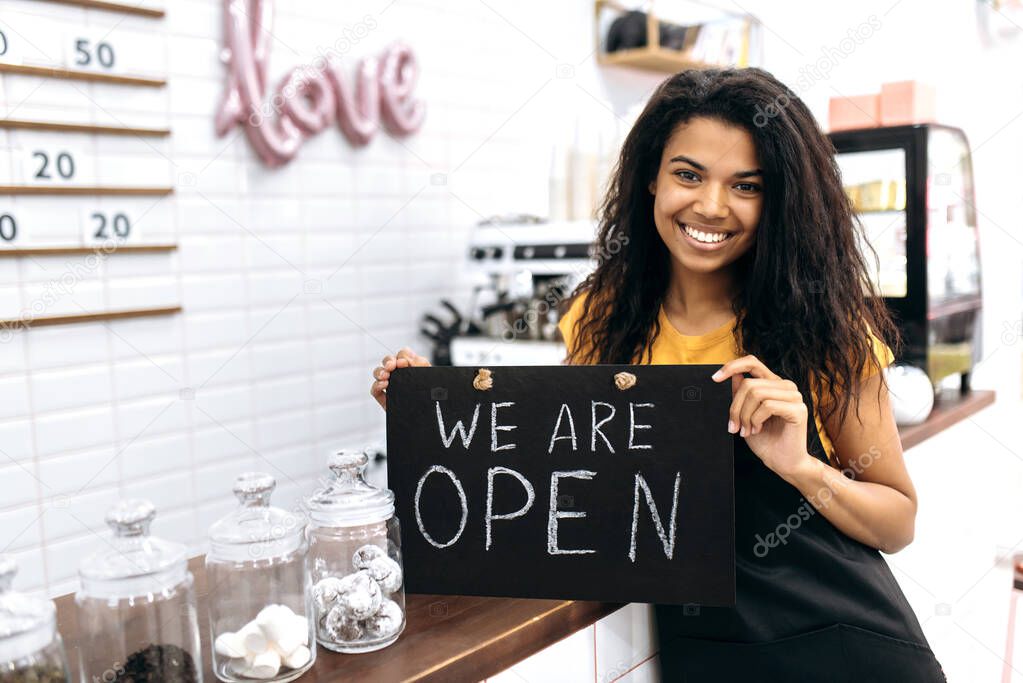 Satisfied African American female barista or waitress, owner of a coffee shop or restaurant, stands holding a sign we are open near the bar counter and friendly smiling