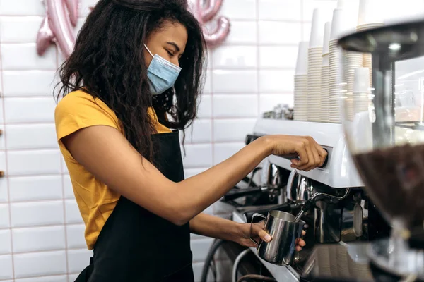 Young African American female barista wearing a medical mask and black apron makes coffee for a coffee shop visitor at their. Owner of small business. Cafe, service industry concept