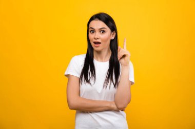 Beyaz, basit tişörtlü, oldukça heyecanlı beyaz esmer bir kadın, yeni bir fikir buldu, izole edilmiş turuncu arka planda duruyor, doğrudan kameraya bakıyor.