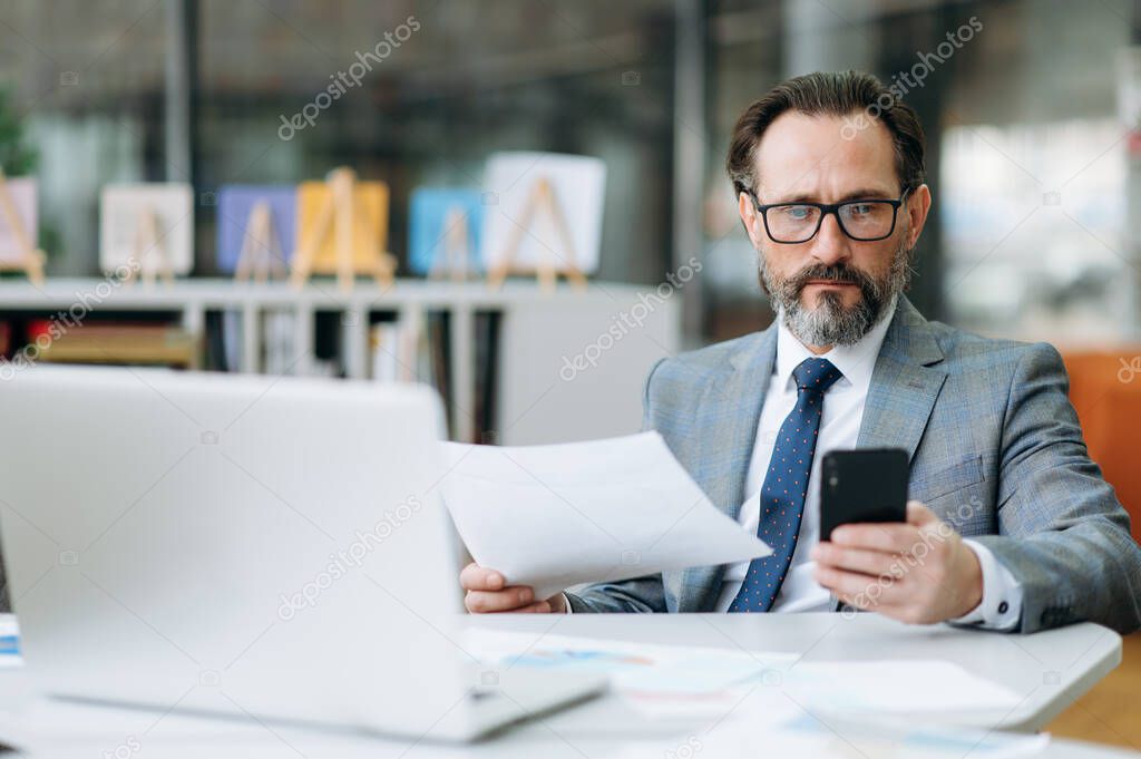 Serious middle aged male employee sits at the table in modern office, works with documents. Concentrated business man is using smartphone, browsing internet, searching ideas for project or startup