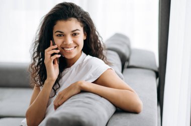 Gülümseyen çok ırklı siyah kıvırcık kız, oturma odasında kanepede oturur ve kameraya bakar, iletişim konsepti