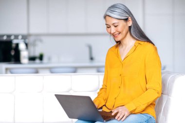 Rahat orta yaşlı, gri saçlı, şık giyinen Asyalı kadın evinde oturuyor, dizüstü bilgisayar kullanıyor, internetten alışveriş yapıyor, evden çalışıyor ya da uzaktan çalışıyor, sosyal ağlara takılıyor.
