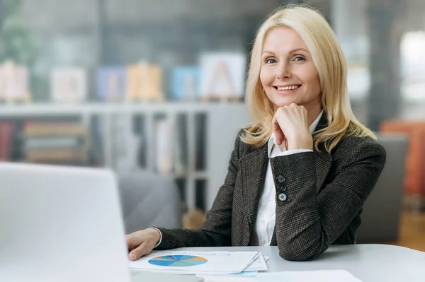 Portrait of successful middle aged business woman, ceo or manager in modern office. Elegant confident blonde lady in stylish formal suit sits at the work desk, look at the camera and friendly smiling