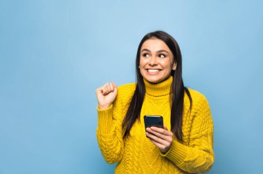 Kafkasyalı mutlu genç bir kadının portresi, akıllı telefonu elinde tutuyor, iyi bir haber alıyor veya beklenmedik bir mesaj alıyor, izole edilmiş mavi arka planda duruyor.