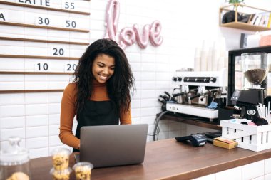 Tatmin olmuş, siyah önlüklü Afro-Amerikan garson, kafeterya sahibi, dizüstü bilgisayarda çalışan, kafede barın arkasında beklerken online siparişleri işleyen.