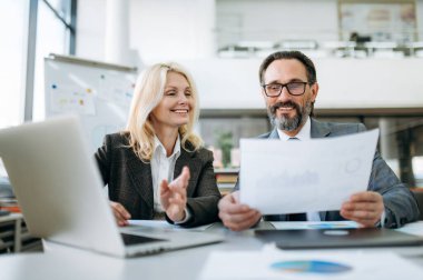 Finansal grafiklere bakarak modern ofiste evrak işleri yapan neşeli ve başarılı iş arkadaşları. Masada oturmuş, yeni iş planı üzerinde çalışan olgun ve şık partnerler.
