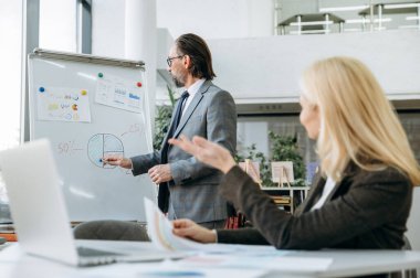 Modern ofiste iş brifing toplantısı kavramı. Şık takım elbiseli kıdemli yetişkin iş adamı, başarılı bayan meslektaşımıza finansal rapor sunacak.