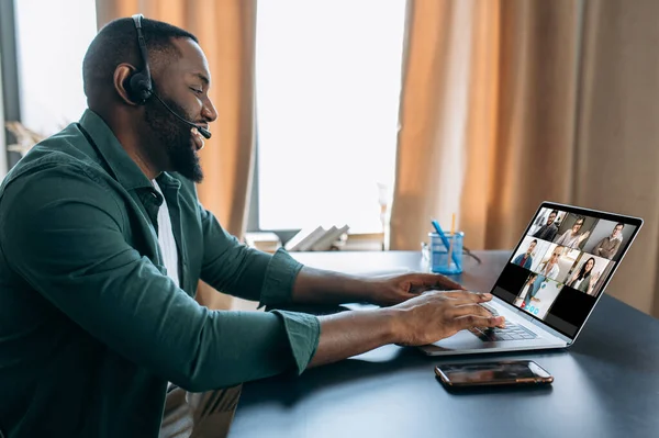 Distance learning, online lesson. African American man studying online, listens to an online lesson via video conference using laptop, on laptop screen multiracial people and teacher, online webinar
