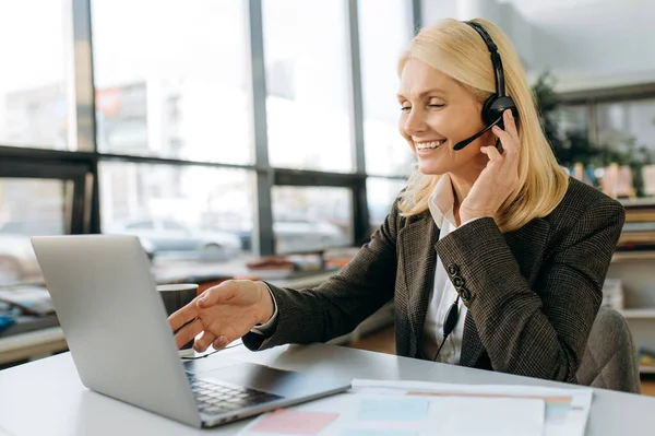 Elegant female employee of support line in headset is using laptop, sitting at desk. Middle aged woman manager conducts online business meeting, communication concept