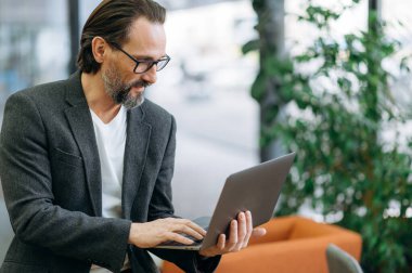 Yoğunlaşmış erkek çalışan dizüstü bilgisayar kullanarak proje üzerinde çalışıyor. Ciddi orta yaşlı işadamı işe odaklandı, başlangıç için bazı fikirleri araştırıyor, internette geziniyor
