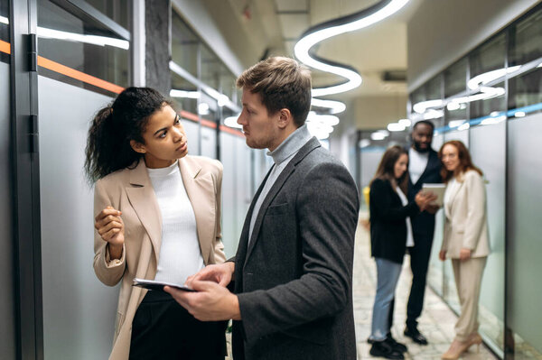 Two multiracial successful colleagues communicate in a modern office, discuss new ideas, think about a plan, in the background their colleagues