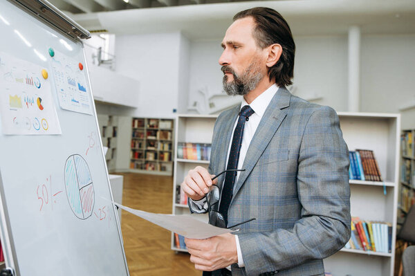 Serious influential businessman is learning financial graphs, standing near flipchart in office. Concentrated senior male developing important project, looks at the marker board