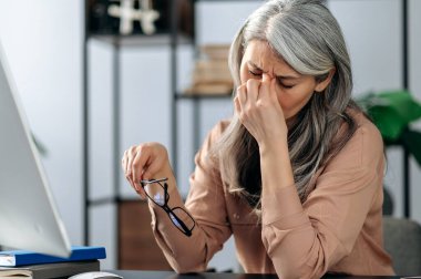 Bilgisayar çalışmasından sonra, elinde gözlük tutan yorgun yaşlı kadın baş ağrısı çekiyor. Üzgün, olgun, gri saçlı, yorgun bir kadın iş yerinde masaj yaptırır.