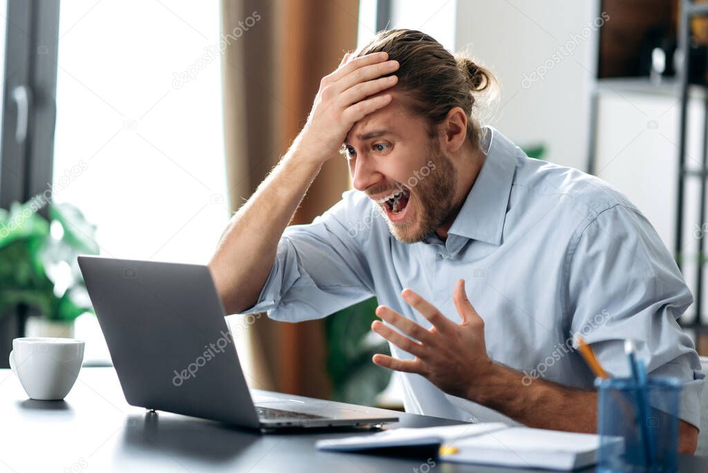 Shocked annoyed caucasian busy guy looking at a laptop in confusion, received unexpected news, failed a deal, shouts, holds his head with his hand, sitting at the work desk