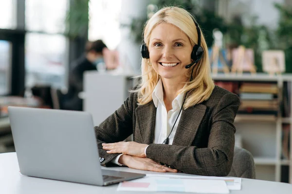 Middle aged woman manager in headset and stylish elegant suit is looking at the camera, smiling. Happy successful female operator of call center is sitting at the desk in office