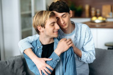 Homoseksüel erkekler. Sevgi dolu eşcinsel çift, evde oturuyor, şefkatle sarılıyor ve birbirlerini okşuyor, oturma odasındaki kanepede birlikte dinleniyorlar.