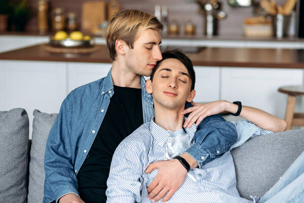 Homosexual guys. Loving gay couple, sitting at home, tenderly hugging and caressing each other, relaxing together on the sofa in the living room. Homosexual relationship concept